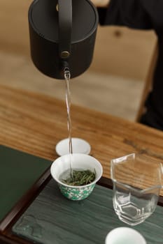 A serene and peaceful tea brewing process featuring a black kettle pouring hot water over fresh green tea leaves