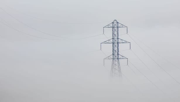 A large metal electricity pylon stands tall amidst a thick fog. Power lines stretch across the misty landscape. No people are visible.