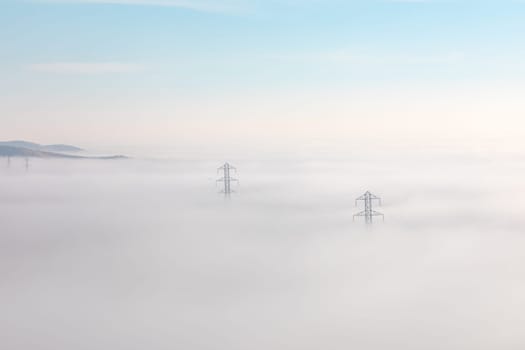 A landscape view of power lines stretching across a thick layer of fog. The fog obscures the terrain, with only the tops of hills visible in the distance
