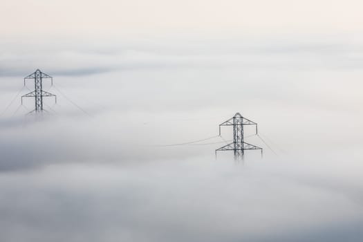Power lines stretch through a thick fog. Metal pylons pierce the misty landscape, carrying electricity unseen. The fog obscures the ground below, highlighting the infrastructure above.