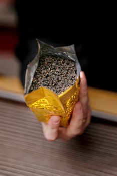 A person holds a golden pouch overflowing with rich, dark tea leaves, highlighting their exquisite aromas