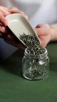 A person carefully pours fragrant green tea leaves into a clear glass jar, showcasing a lovely tea preparation moment