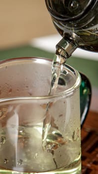 A closeup shot is capturing the moment of hot tea being poured from a kettle into a delicate glass mug