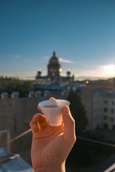 A hand holds a small cup, enjoying the stunning sunsetlit city skyline, creating an unforgettable moment