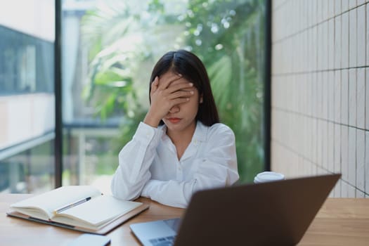Stressed young woman in a modern office setting, covering her face with one hand and surrounded by a laptop and notebook, reflecting the challenges of work and deadlines.