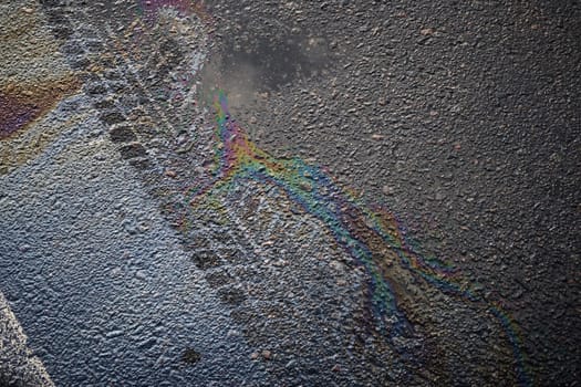 Close-up of tire marks and gasoline or oil spill creating a rainbow effect on wet asphalt road surface.