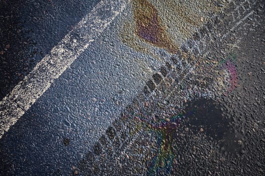 Gasoline rainbow spill reflecting on wet asphalt road surface with white stripe.