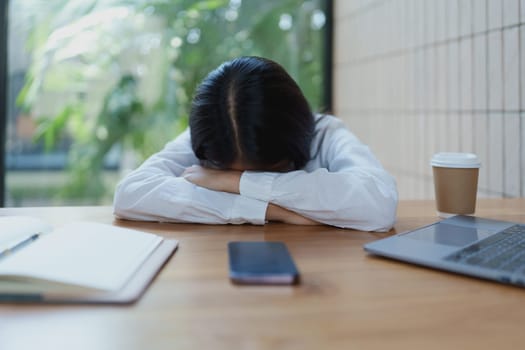 A young woman sits at her desk, head resting on her arms, expressing stress and fatigue in a serene workspace filled with natural light and greenery.