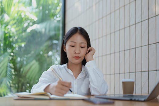A focused woman writes in her notebook at a cozy wooden table, enjoying a coffee while surrounded by a serene and bright atmosphere filled with greenery.