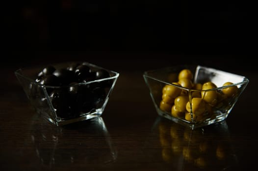 Two glass bowls filled with black and green olives on a reflective wooden surface, showcasing a contrast in color and texture.