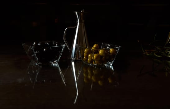 Dark, moody setup featuring black and green olives in glass bowls beside a silver oil can.