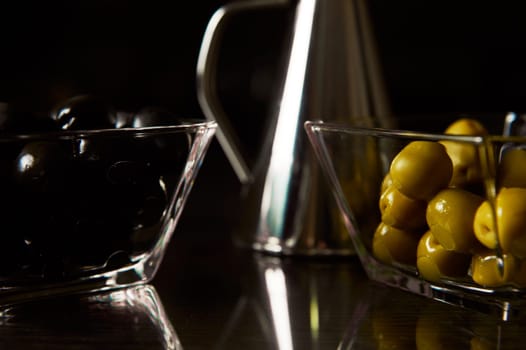 Close-up of black and green olives in glass dishes, focusing on textures and reflections with a metal container in the background creating a modern culinary aesthetic.