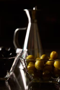Close-up of glass bowls containing green and black olives, paired with a silver oil cruet, under dramatic lighting.
