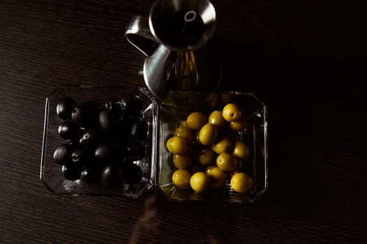 Close-up of black and green olives in clear glass bowls beside a shiny silver oil jug on a dark wooden surface.