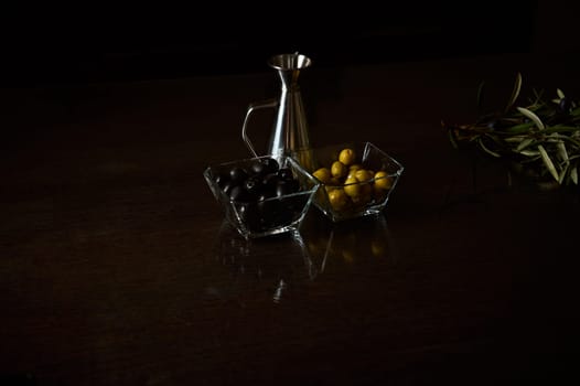 A sophisticated arrangement featuring black and green olives in glass bowls with a metal oil decanter on a dark wooden surface.