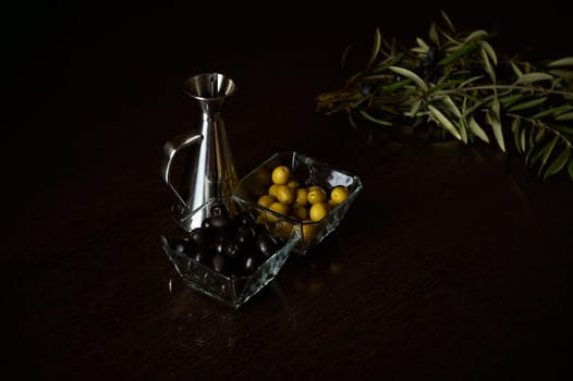 Fresh olive branches next to glass bowls of olives and a metal oil jug on a dark table.
