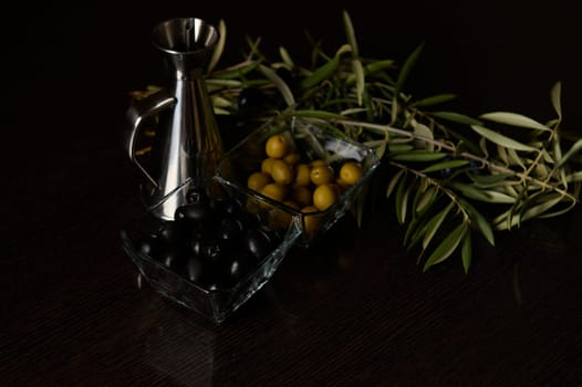 A sophisticated display featuring black and green olives, an olive sprig, and a metallic oil dispenser on a dark surface.