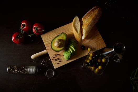 Top view of fresh ingredients including avocado, tomatoes, olives, bread, and spices on a wooden board.