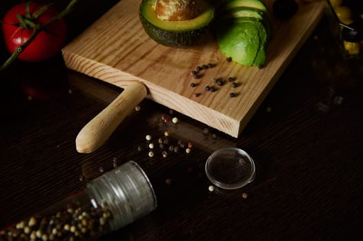 A rustic wooden cutting board with fresh avocado slices, surrounded by peppercorns and a tomato, on a dark kitchen countertop.