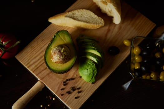 A rustic presentation of fresh sliced avocado with bread and assorted olives on a wooden board.