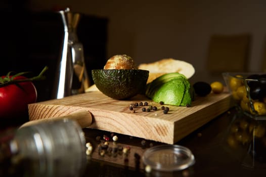 Sliced avocado on a chopping board with spices, olives, and tomatoes. Rustic kitchen setting conveying freshness and culinary creativity.