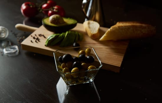 A selection of fresh olives and avocado slices on a cutting board with bread and tomatoes. Prepared for a healthy snack or appetizer on a dark, moody kitchen surface.