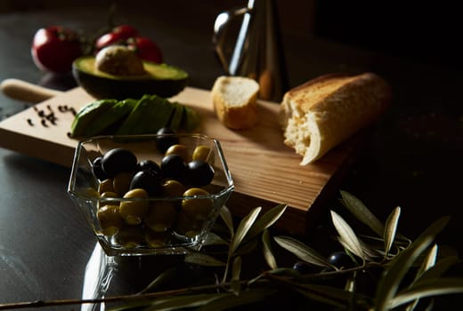 A vibrant display of fresh Mediterranean ingredients including olives, avocado, bread, and tomatoes. The warm, rustic setting highlights a wooden cutting board and a bowl of olives.