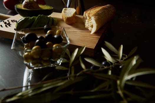 A rustic composition featuring fresh olives, sliced avocado, and baguette, evoking Mediterranean flavors.