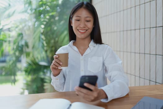 A cheerful young woman sits at a wooden table, sipping coffee and using her smartphone. The setting is bright and filled with greenery, reflecting a modern, relaxed lifestyle.
