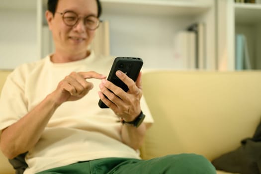 Happy elderly Asian man in eyeglasses using smartphone on couch at home.