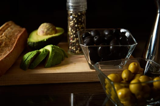 A selection of fresh ingredients including avocado, olives, and bread are artfully displayed on a wooden board, evoking a Mediterranean culinary theme.