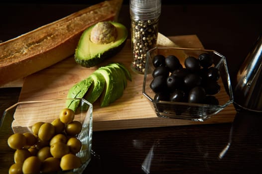 A variety of fresh ingredients including sliced avocado, green and black olives, and bread, displayed on a cutting board for a healthy meal preparation.