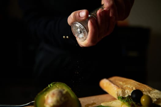 A person seasoning fresh avocado with salt, highlighting kitchen preparation. The scene includes bread and olives, emphasizing culinary art and home cooking atmosphere.
