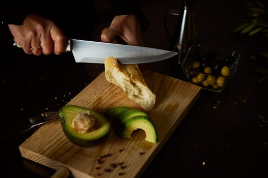 A person slices bread on a wooden board with avocado and olives nearby, creating a culinary scene.