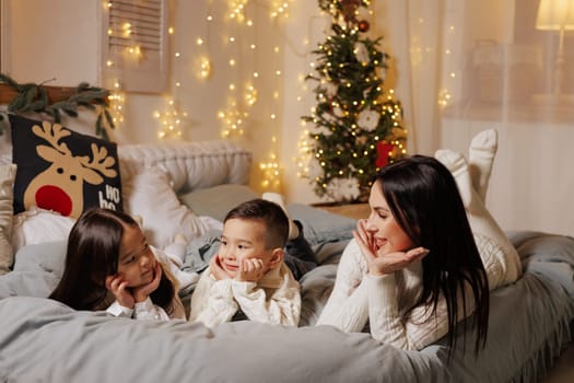 mother and two children lying on bed on christmas happy evening in cozy room decorated with garlands,
