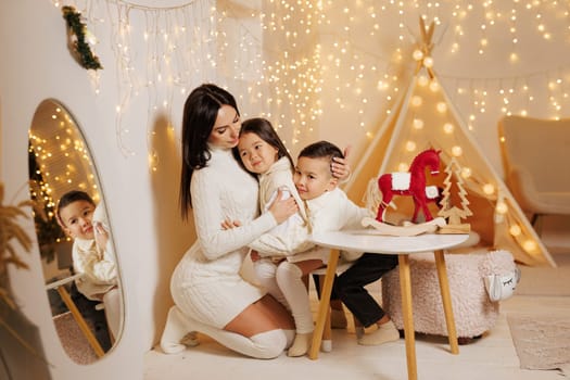 two children hugging mother on christmas evening in cozy room decorated with new year garlands, happy family on christmas