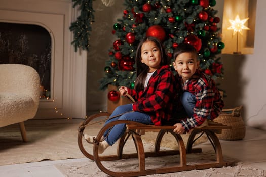 Two children sitting on a wooden sled in a cozy, festive living room decorated for Christmas. A Christmas tree adorned with red and green ornaments is in the background, creating a warm holiday atmosphere