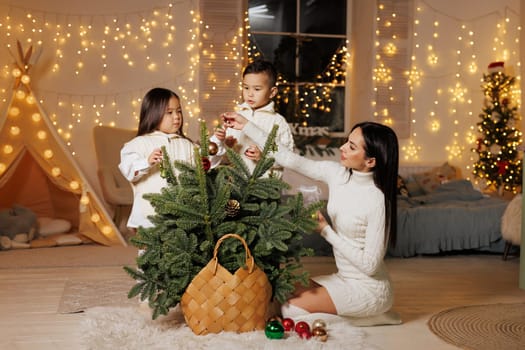 mother and two children decorate a small Christmas tree on Christmas evening in a cozy room decorated with New Year garlands, happy family at Christmas,