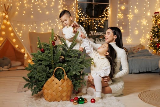 A mother and her two children decorating a small Christmas tree in a cozy, festive room adorned with fairy lights. The children are playfully reaching for ornaments, while the mother watches with a smile. The atmosphere is warm and joyful,