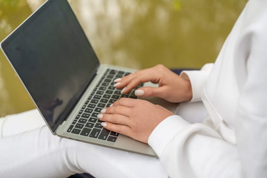 A woman is typing on a laptop while sitting on a chair. She is wearing a white shirt and white pants. The laptop is open and the screen is black. The woman is focused on her work