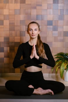 An athletic girl in black sportswear sits in lotus pose at a yoga class in a fitness room among green indoor plants.
