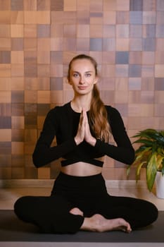 An athletic girl in black sportswear sits in lotus pose at a yoga class in a fitness room among green indoor plants.