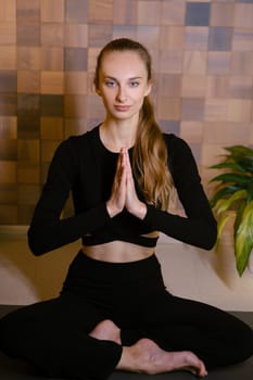An athletic girl in black sportswear sits in lotus pose at a yoga class in a fitness room among green indoor plants.