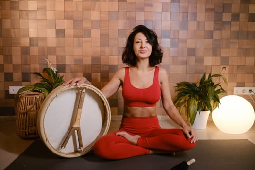 A woman in a red tracksuit sits with a drum in a gymnasium. Kalimeditation class.