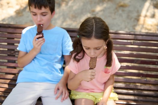 Two kids sitting on a bench savor chocolate ice cream bars outdoors on a sunny day.