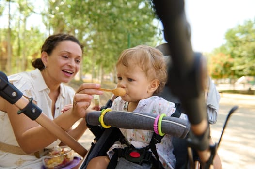 A joyful mother feeding her toddler in a stroller in the park, celebrating outdoor family moments.