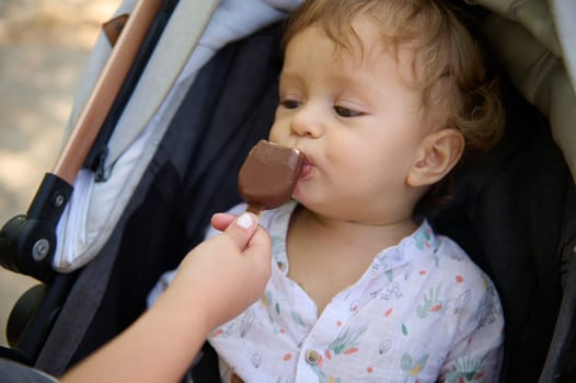 Cute baby sitting in a stroller, having a first taste of chocolate ice cream on a sunny day.