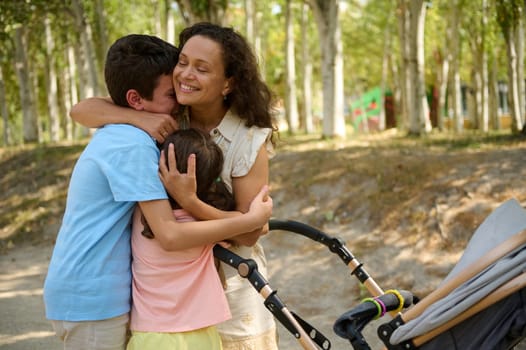 A loving mother hugs her children near a stroller under trees in a sunny park, expressing warmth and closeness.