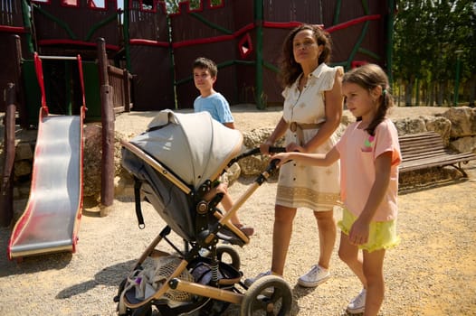 A family spends quality time at the park, exploring and playing together. The mother pushes a stroller while the kids explore nearby play equipment, capturing moments of fun and togetherness.