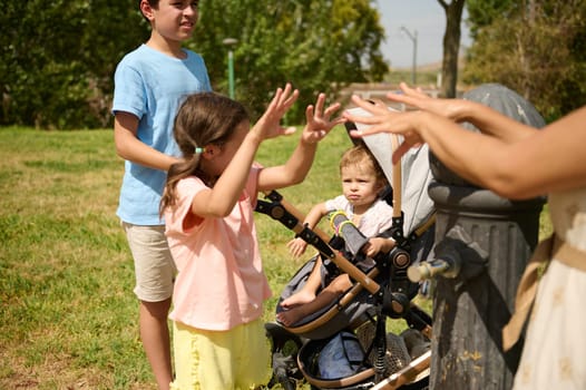 A group of children interacts near a stroller by a water fountain in a sunny park, enjoying playful outdoor activities under the bright sun.
